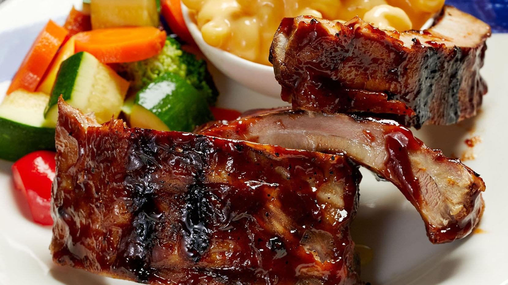 A plate featuring kid-sized portions of glazed ribs, fresh vegetables, and macaroni and cheese.