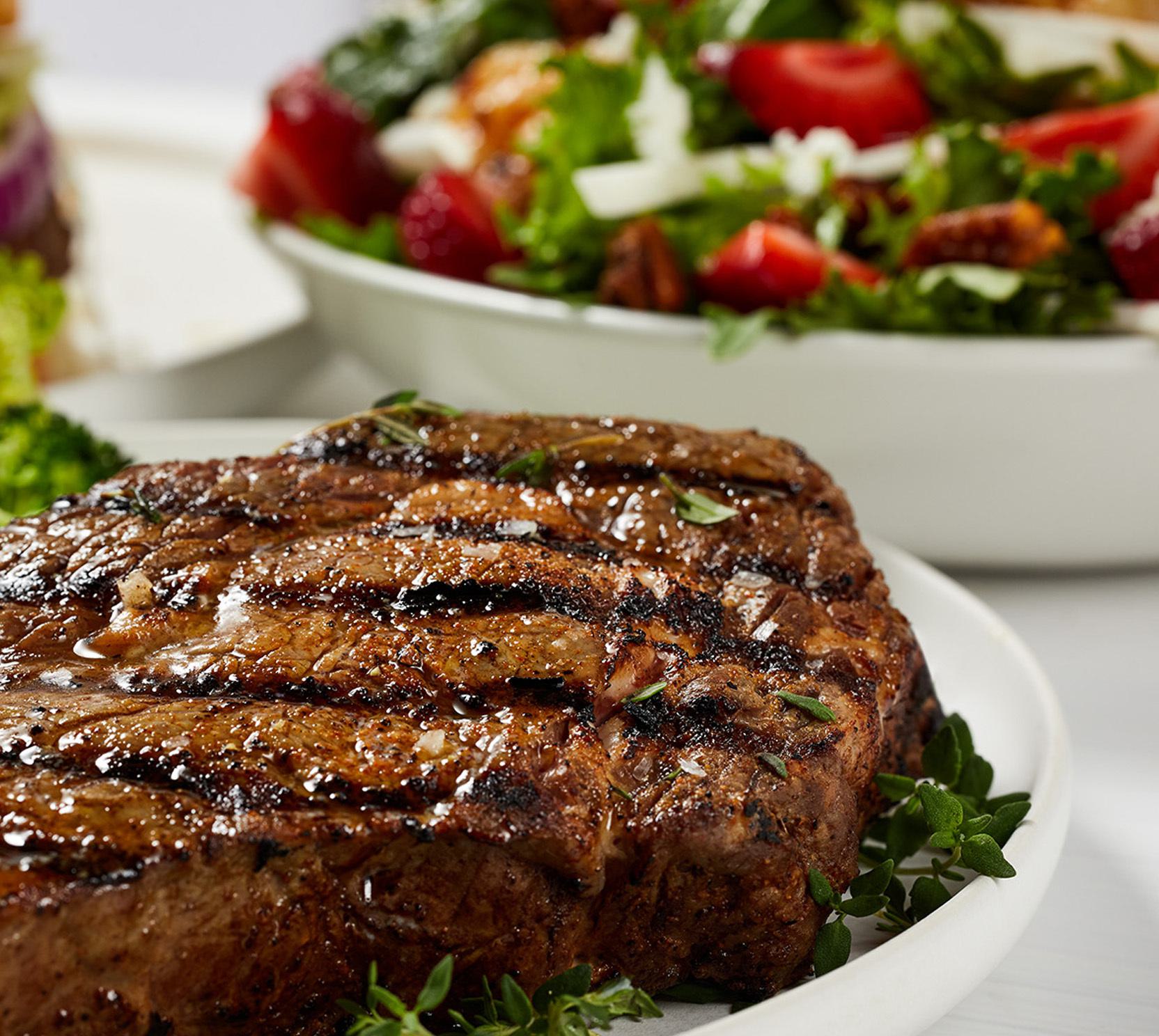 A delicious, steak on a plate, with a salad in the background.